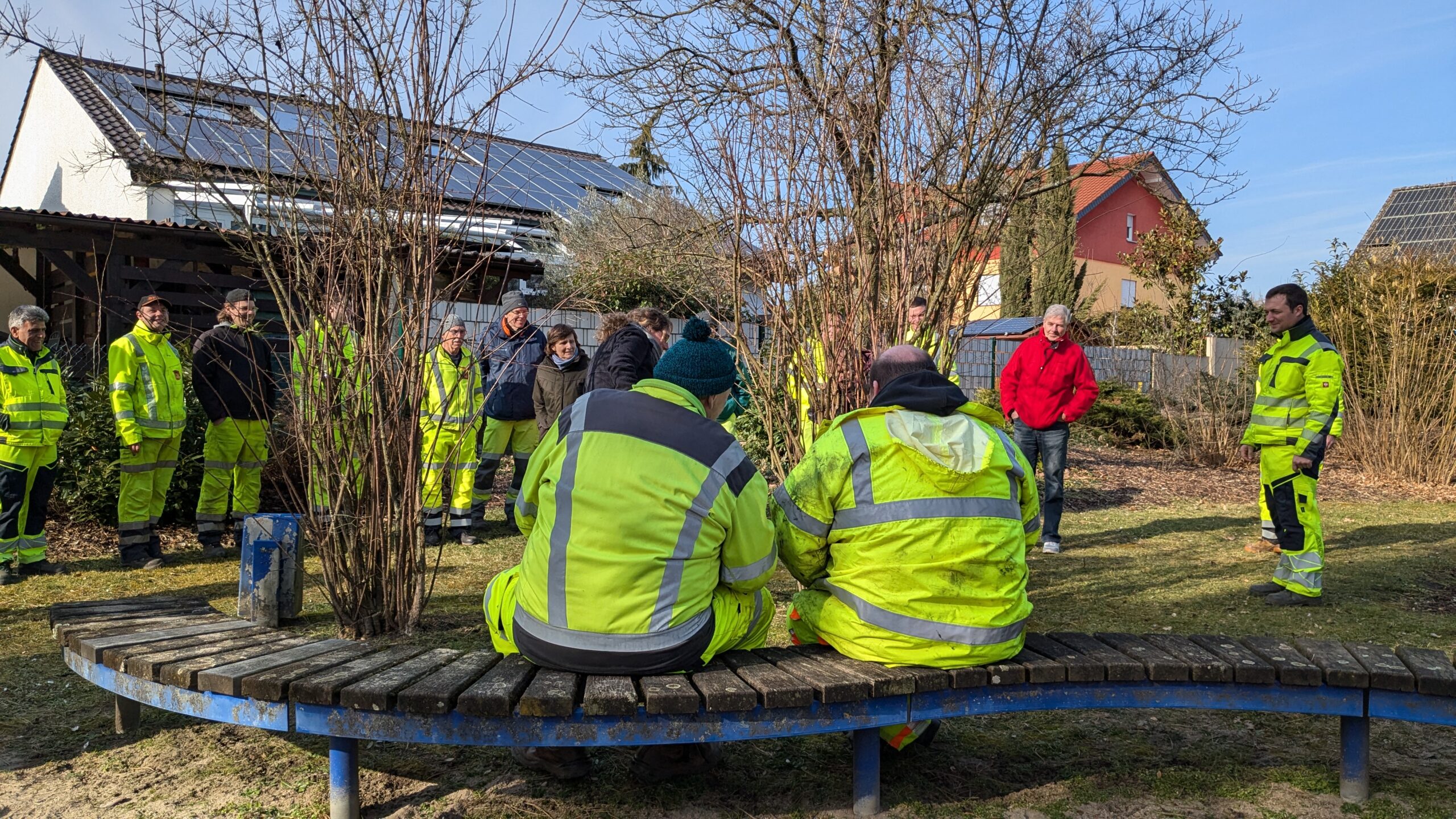 Zwei Personen in Arbeitkleidung schauen Kollegen beim Heckenschnitt zu.