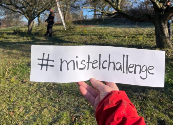 Hand hält Schild mit #mistelchallenge, im Hintergrund Bäume