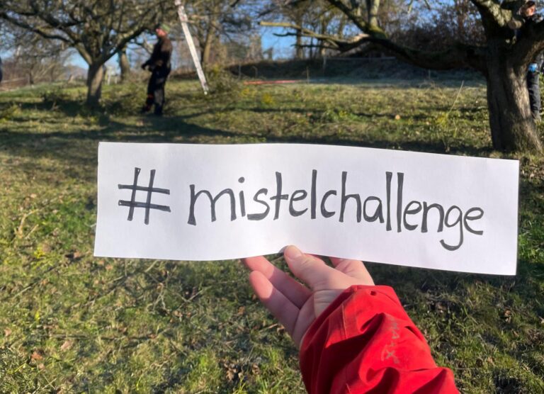 Hand hält Schild mit #mistelchallenge, im Hintergrund Bäume
